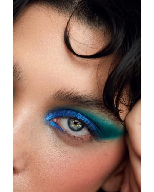 Extreme close-up of a young woman's face shows off her eye, framed by curly dark hair and vibrant blue and green eyeshadow makeup.