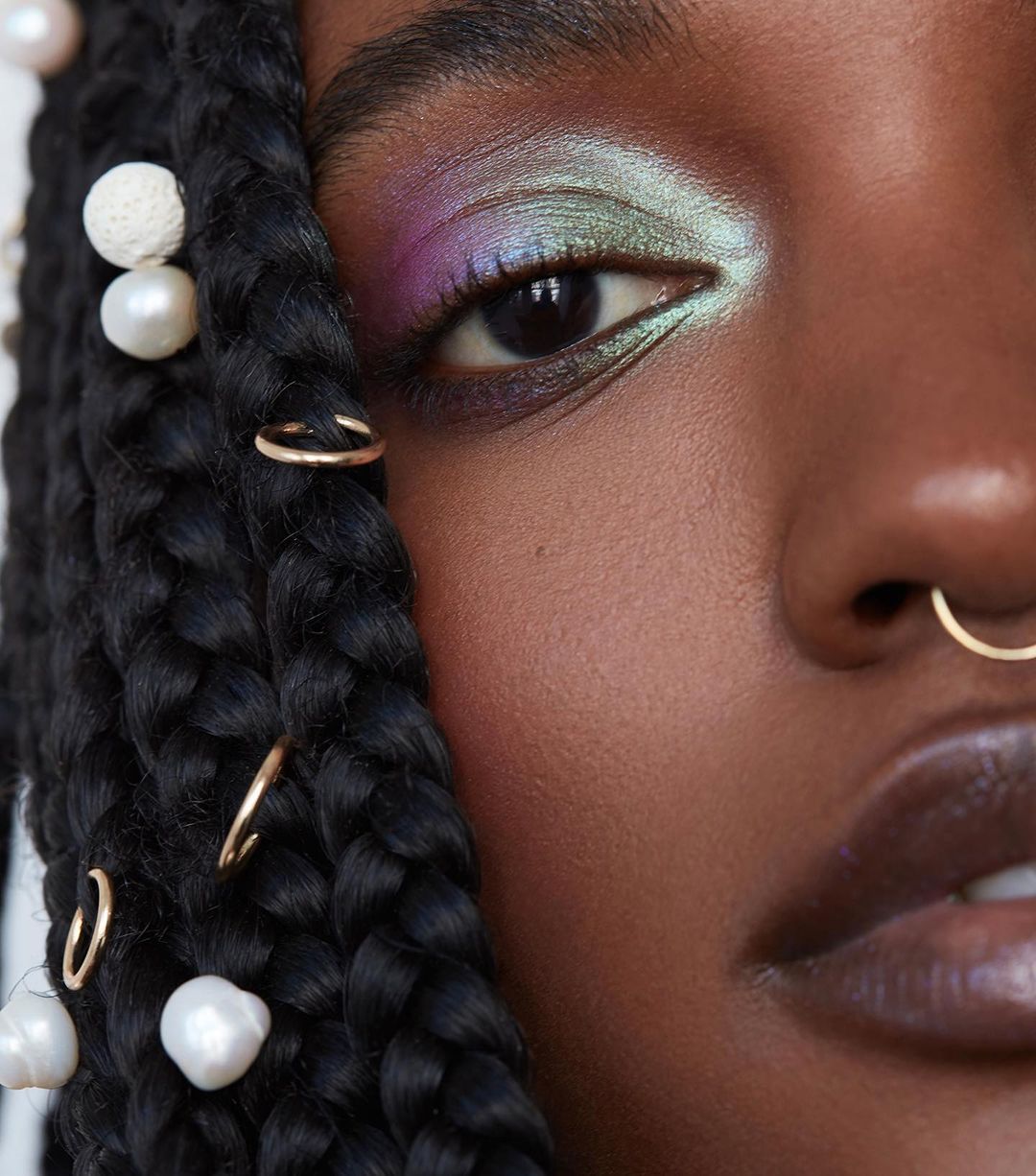 Close-up of a black woman's face showcasing vibrant eyeshadow, pearl hair accessories, and gold nose and braid rings in a beauty shot.
