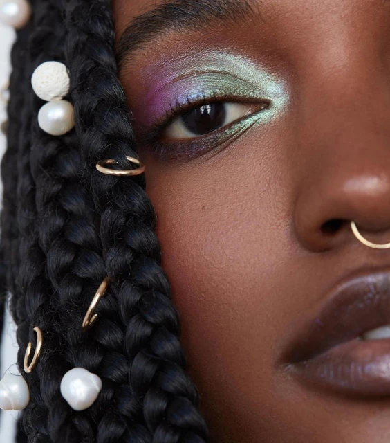 Close-up of a black woman's face showcasing vibrant eyeshadow, pearl hair accessories, and gold nose and braid rings in a beauty shot.
