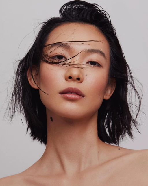 Close-up studio portrait of a young Asian woman with black hair, fair skin, and neutral expression against a neutral grey background.
