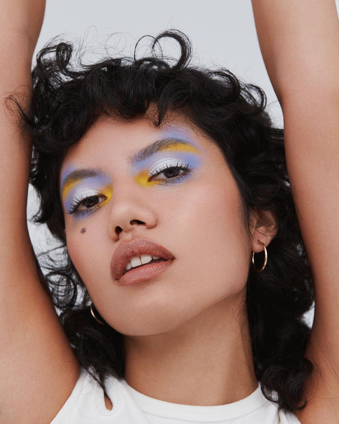 Close-up studio shot of an adult woman with creative yellow, white, and purple makeup on her eyes, and gold hoop earrings.