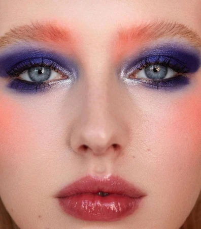 Close-up portrait of a woman with vibrant purple eyeshadow, peach-tinted brows, and glossy pink lips.