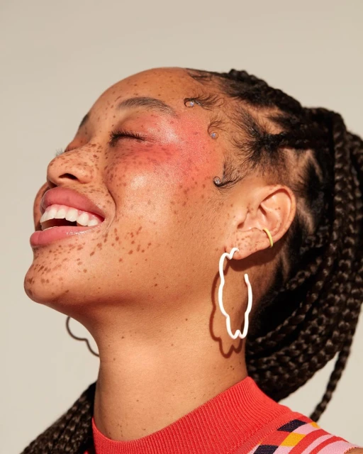 Close-up of a smiling African American woman with freckles, braids, white earrings, and a red patterned top in a studio setting.