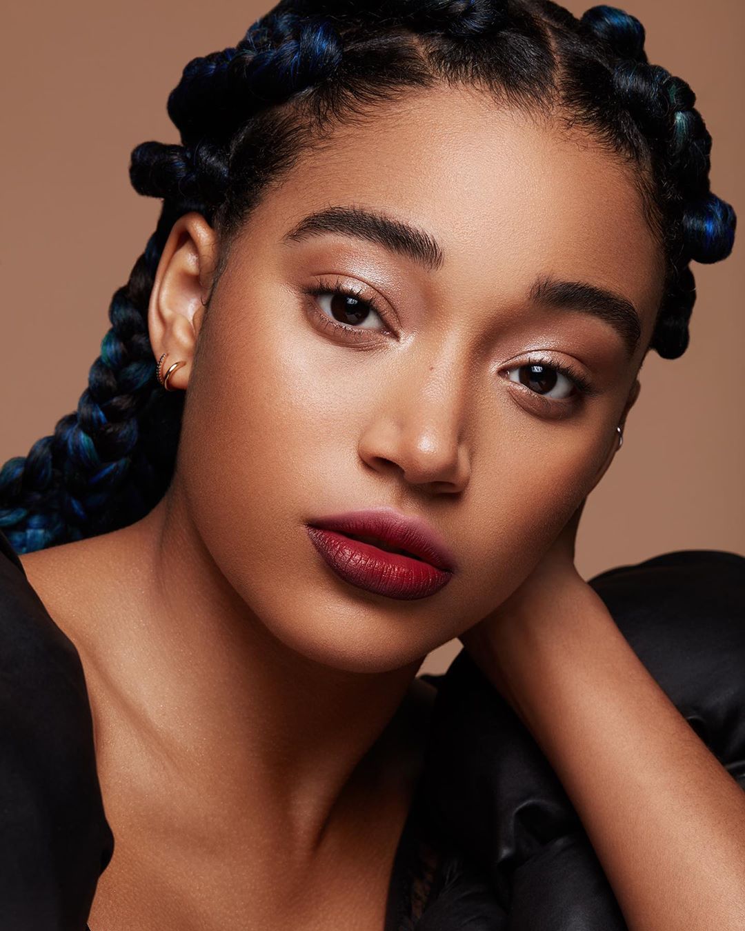 Close-up portrait of a dark-skinned woman with blue-streaked braids and dark red lipstick against a neutral background in a beauty shot.