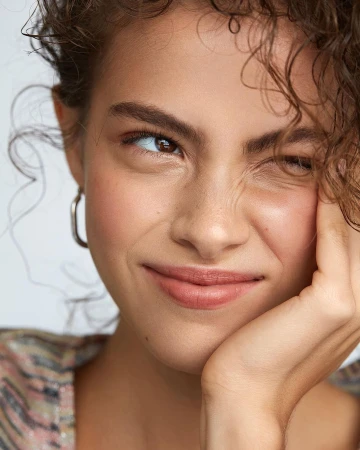 Close-up portrait of a woman with curly brown hair scrunching her face and resting her chin on her hand against a white backdrop.
