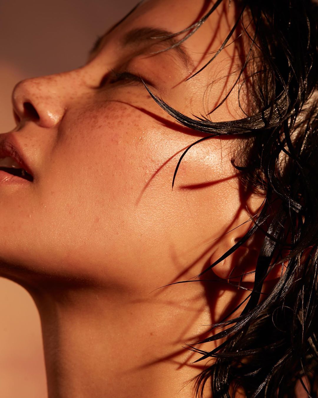 Close-up portrait of a young adult with wet hair, upward gaze, freckles, and warm lighting against a blurred orange backdrop.