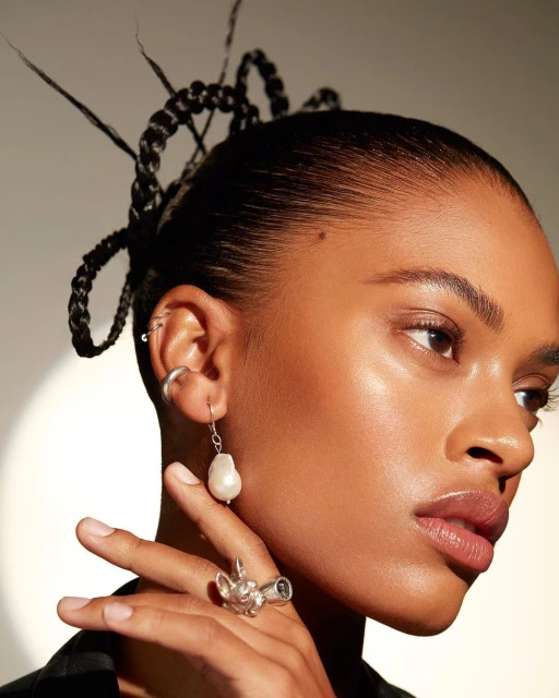 Close-up of a Black woman wearing pearl drop earring and decorative ring. She has black braided hair and neutral makeup.