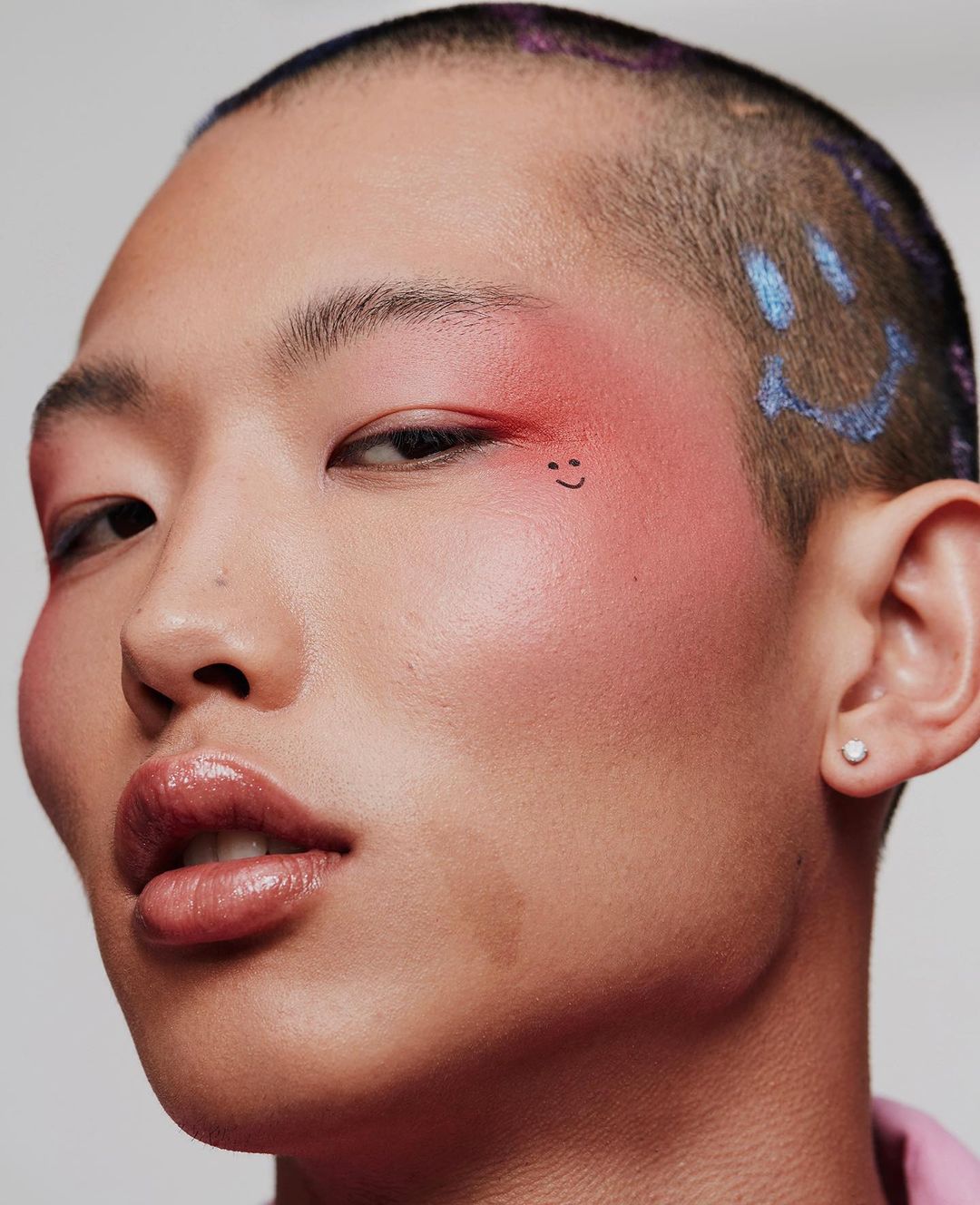 Close-up portrait of an adult person with short hair and makeup, featuring creative face art and a bright, neutral backdrop.