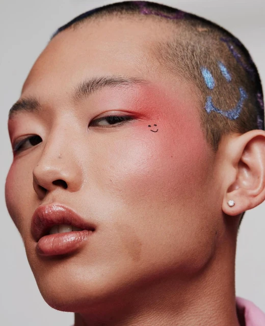 Close-up portrait of an adult person with short hair and makeup, featuring creative face art and a bright, neutral backdrop.