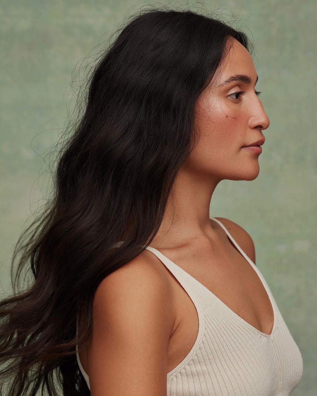 Profile portrait of a brunette woman with long dark wavy hair wearing a white ribbed tank top against a textured green backdrop