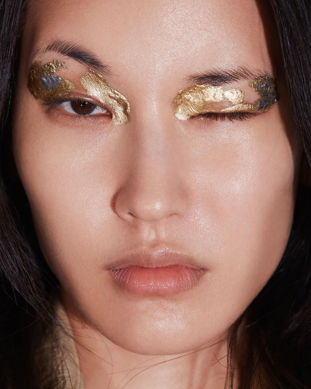 Close-up of an Asian adult female face with dramatic gold foil eye makeup, posing with one eye closed in a beauty or fashion context.