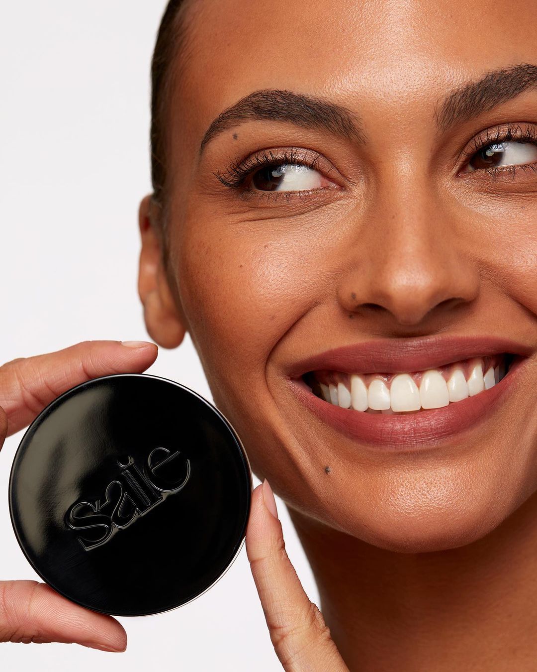 Close-up of a woman holding a black compact with a white logo, set against a clean white background in a beauty or fashion context.