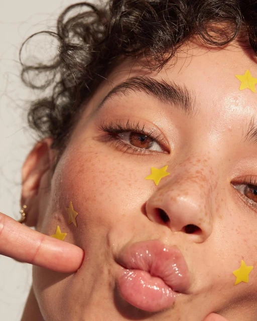 Close-up portrait of an adult woman with fair skin and freckles, puckered lips, and yellow star-shaped face gems on a neutral background.