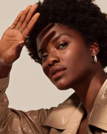 Close-up portrait of a Black woman with natural hair shielding her eyes with her hand, wearing a tan trench coat, and silver hoop earrings.