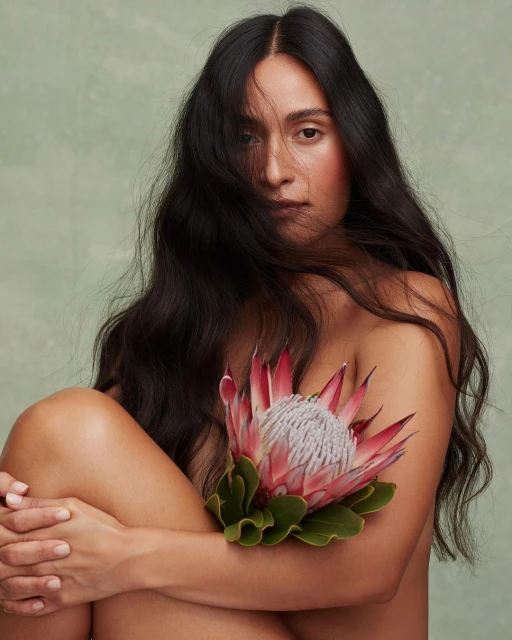 Adult woman with long black hair hugs her knees and holds a pink king protea flower against a mottled light green background.