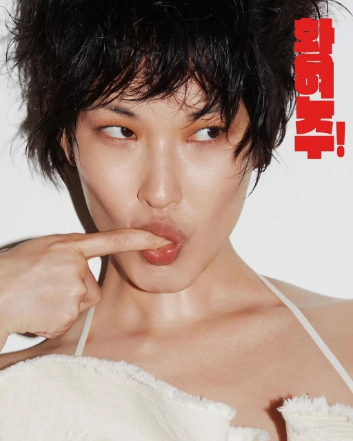 Close-up studio portrait of a young woman with short black hair, light skin, and a white top, with her finger touching her lip.