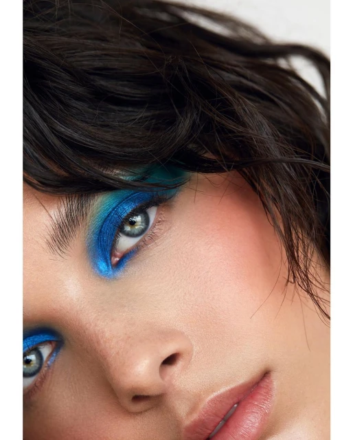 Close-up beauty shot of a young adult woman with dark hair and bright blue eye shadow, fair skin, in a brightly lit photo studio.