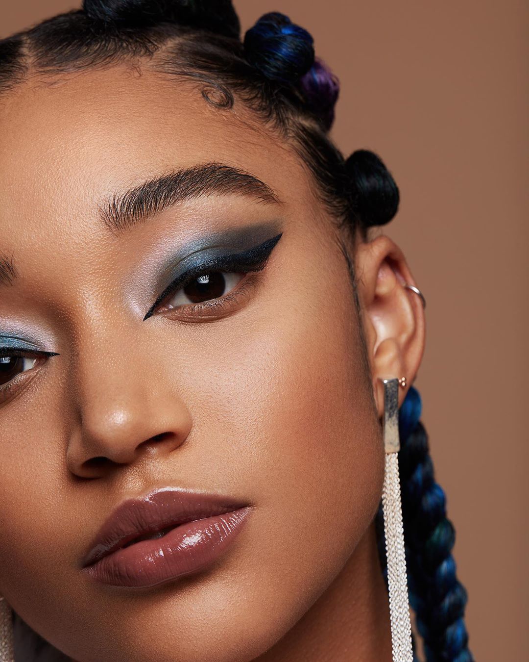 Close-up portrait of a young woman with blue braided hair, creative makeup, and silver earrings, set against a neutral backdrop.