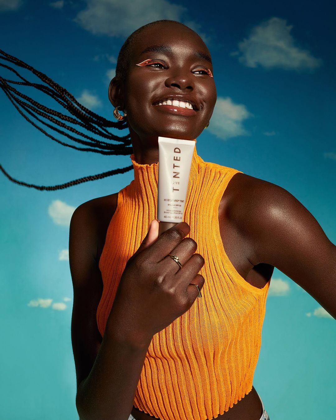 Smiling Black woman holds a white tinted sunscreen tube against a blue sky backdrop; model with orange eyeshadow and braided hair.