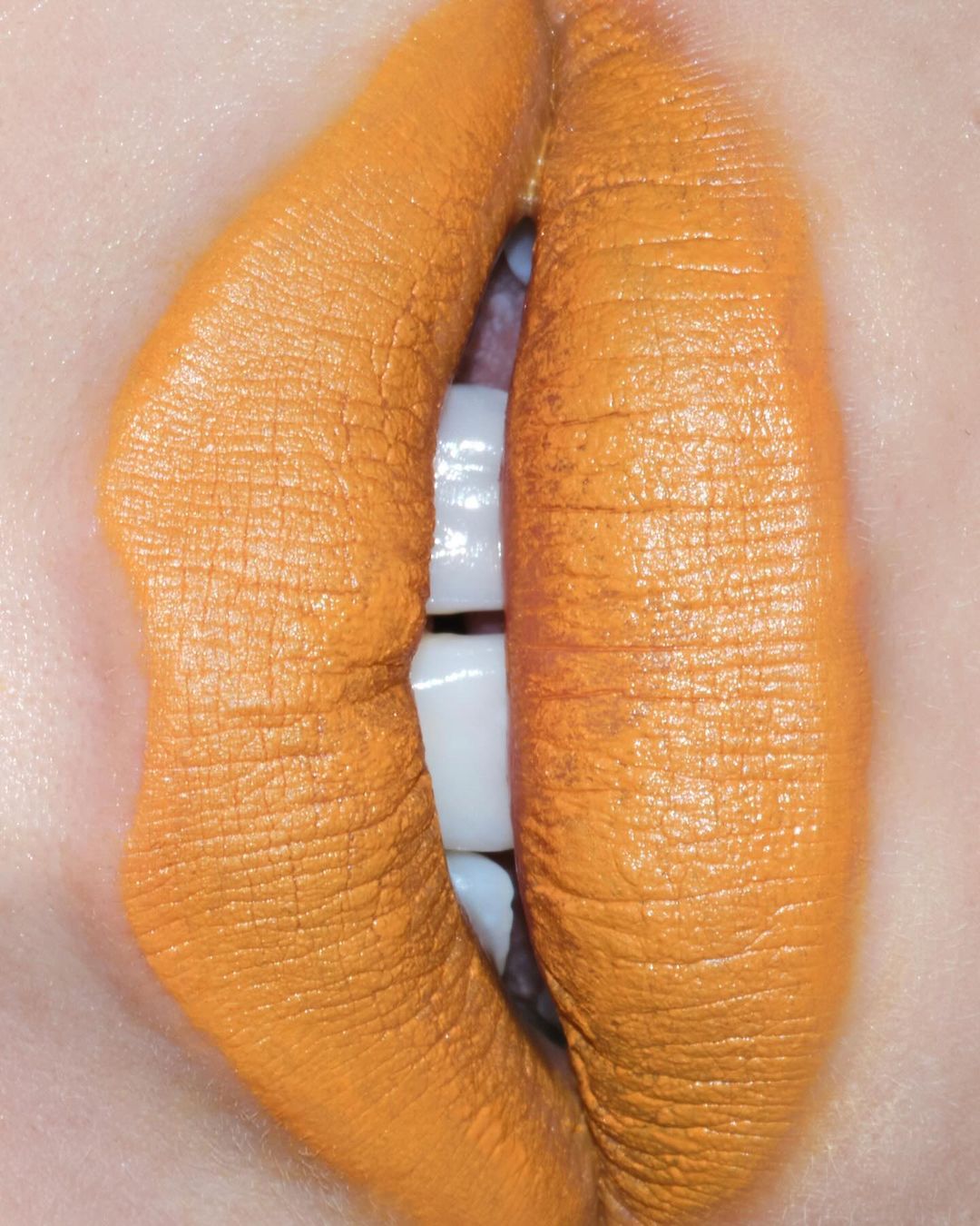 Close-up macro shot of lips wearing matte orange lipstick, revealing white teeth through a slight gap.