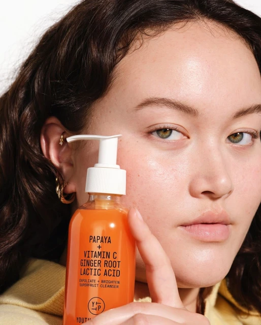 A young adult female model is shown indoors holding and displaying a bottle of youth boosting skin cleanser against a white background.