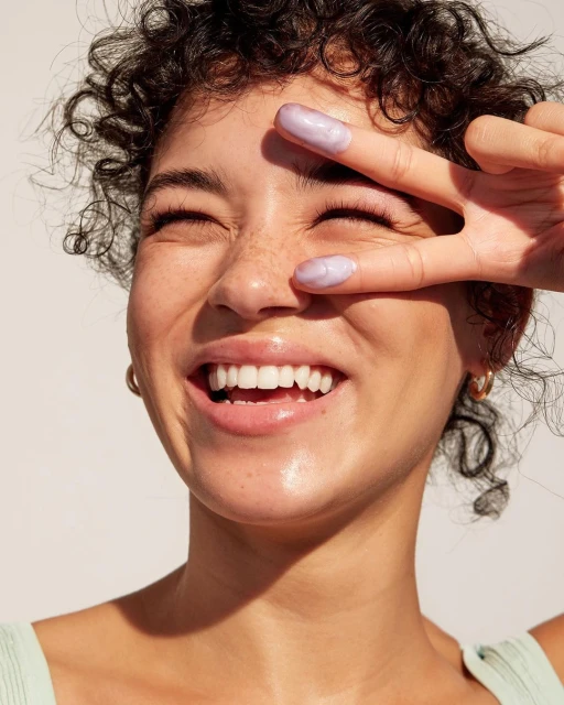 Close-up of a smiling woman with curly hair making a peace sign near her eye. She is wearing a light green shirt and gold earrings.