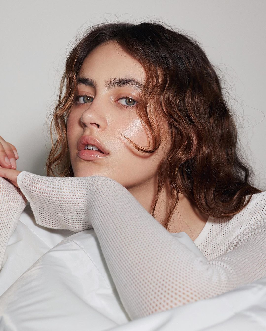 Close-up portrait of a young woman with wavy brown hair, fair skin, and a natural expression, wearing a white knitted top, against a plain background.