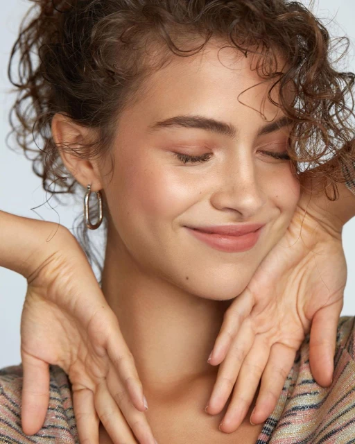Close-up portrait of a young woman with curly brown hair and light skin, eyes closed, wearing hoop earrings and a striped shirt.