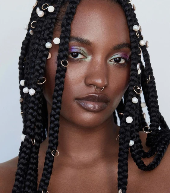Close-up portrait of a dark-skinned woman with box braids, adorned with pearls and gold rings, against a clean white background.