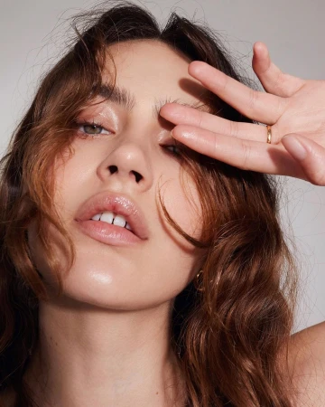 Close-up portrait of a woman holding her hand near her face with glossy lips and wavy brown hair against a neutral background.