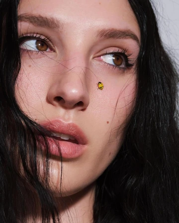 Close-up portrait of a fair-skinned woman with brunette hair and brown eyes looking up, featuring a yellow ladybug on her cheek.