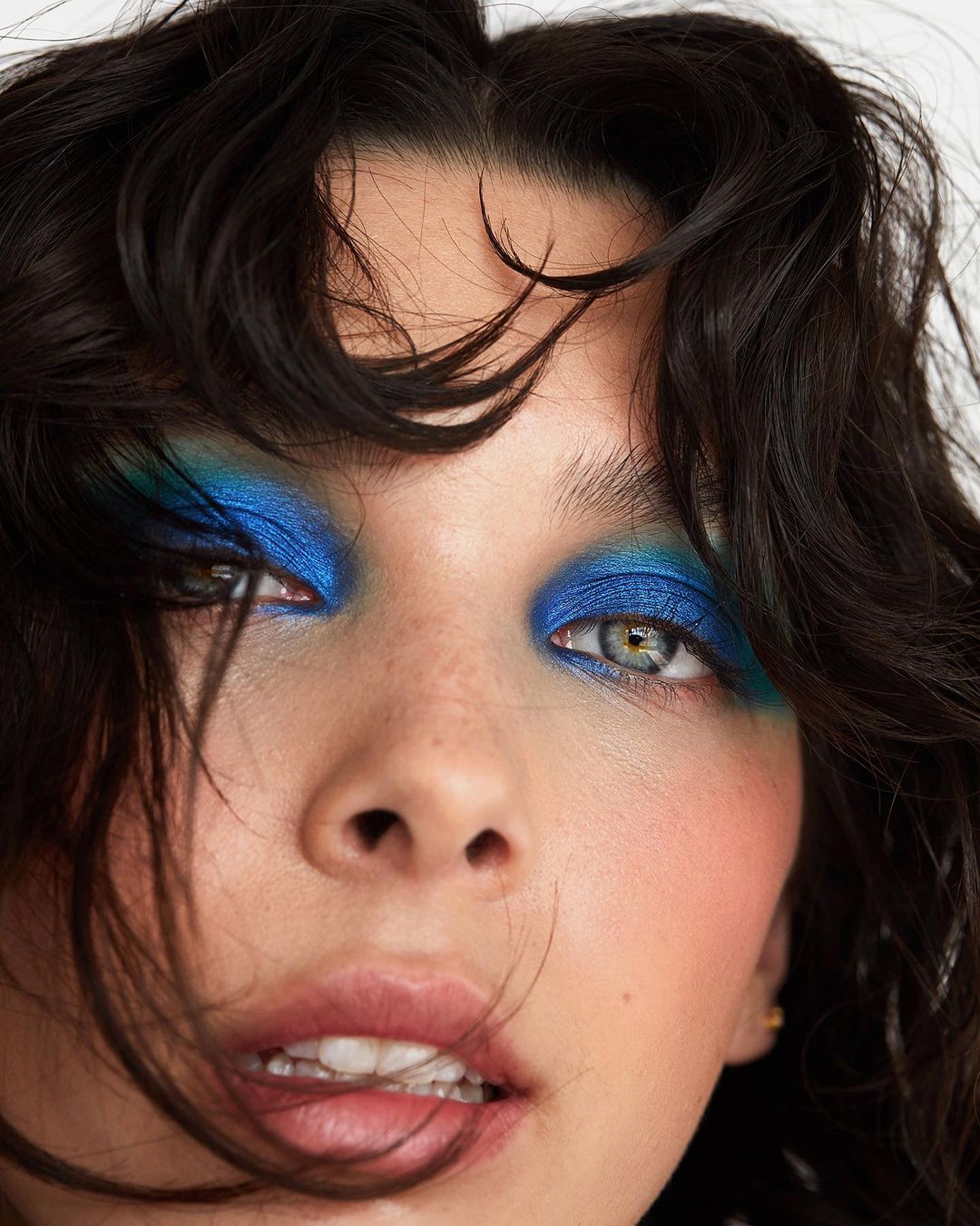 Close-up portrait of a woman with vibrant blue eyeshadow and dark tousled hair framing her face against a plain background
