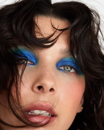Close-up portrait of a woman with vibrant blue eyeshadow and dark tousled hair framing her face against a plain background