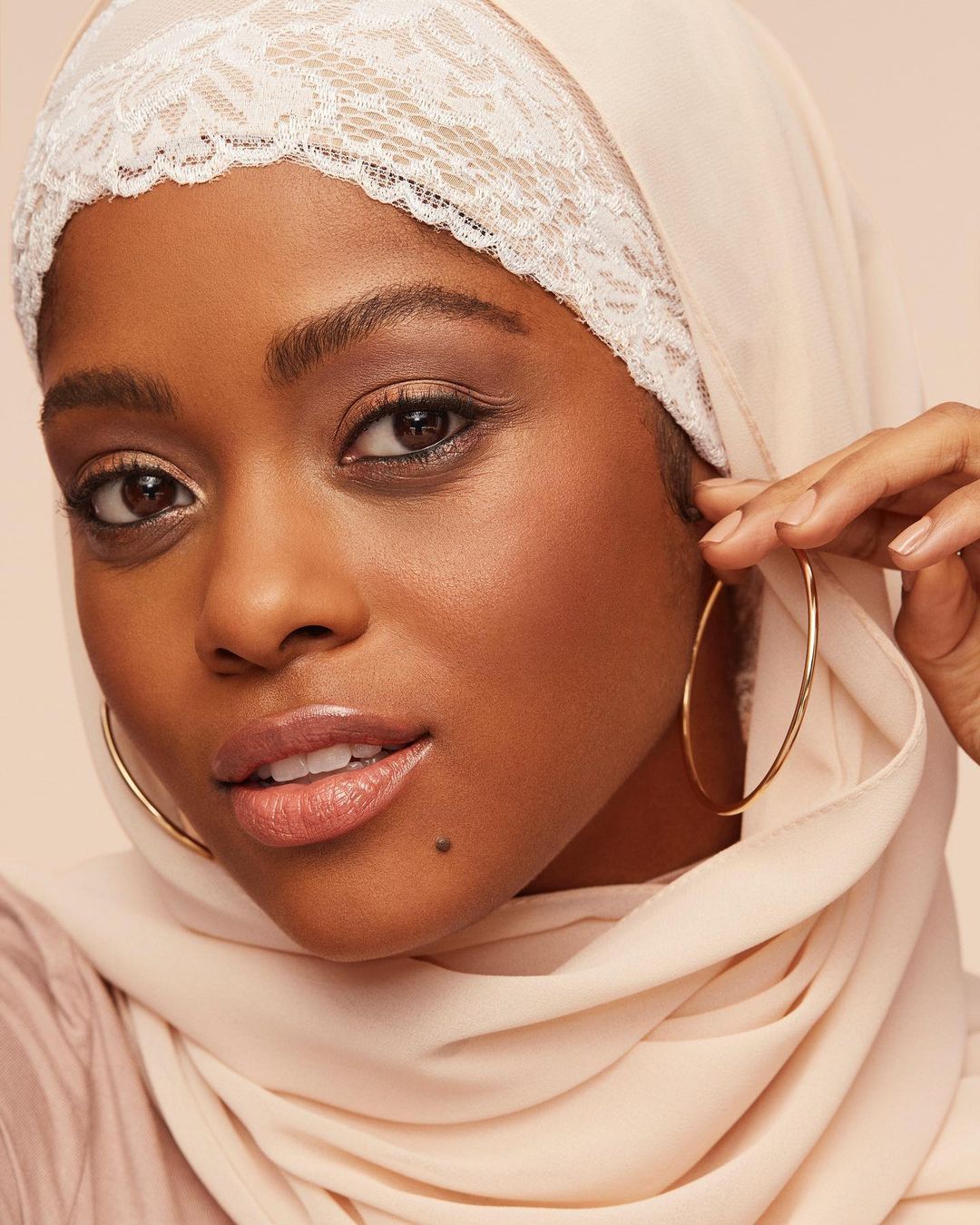 Close-up portrait of an African-American woman wearing a cream-colored hijab and large gold hoop earrings against a light background.