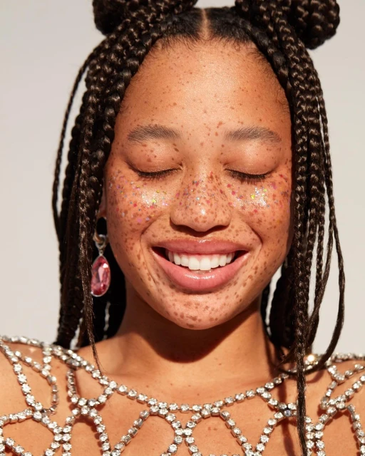 Close-up of a smiling woman with freckles, braided space buns, rhinestone top, and tear-drop earrings against a neutral background.