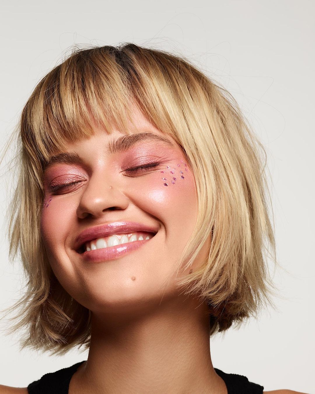 Smiling young woman with blonde bob haircut and pink glittery makeup in a close-up studio shot on a white backdrop.