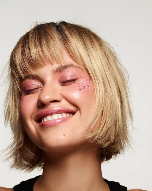 Smiling young woman with blonde bob haircut and pink glittery makeup in a close-up studio shot on a white backdrop.