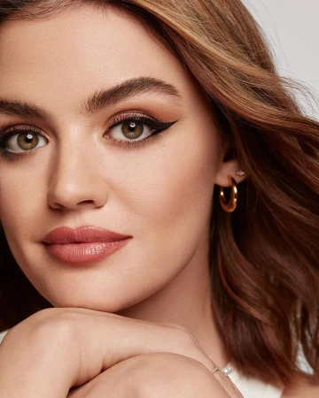 Close-up beauty portrait of a woman with dramatic winged eyeliner, glowing skin, and gold hoop earrings against a neutral background