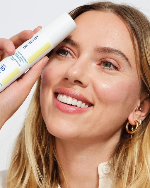 Blonde adult woman with fair skin smiles in a medium shot holding a white cosmetic bottle of SPF 30 mineral sunscreen.