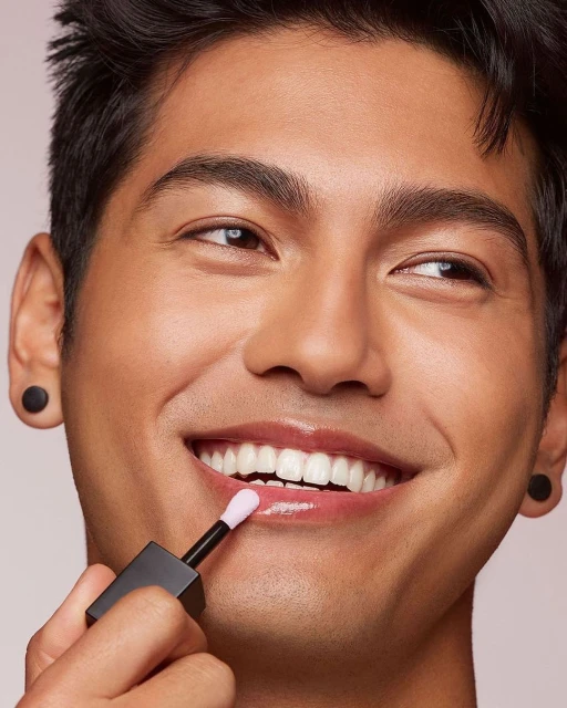 Close-up of a man applying lip gloss with applicator brush to his lips, smiling, against a light neutral background, beauty shot.