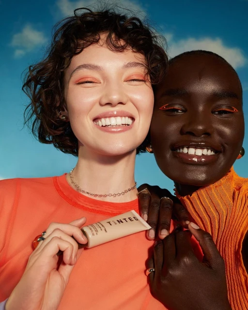 Two smiling women pose with a tinted moisturizer against a blue, slightly cloudy sky. One woman holds the product.