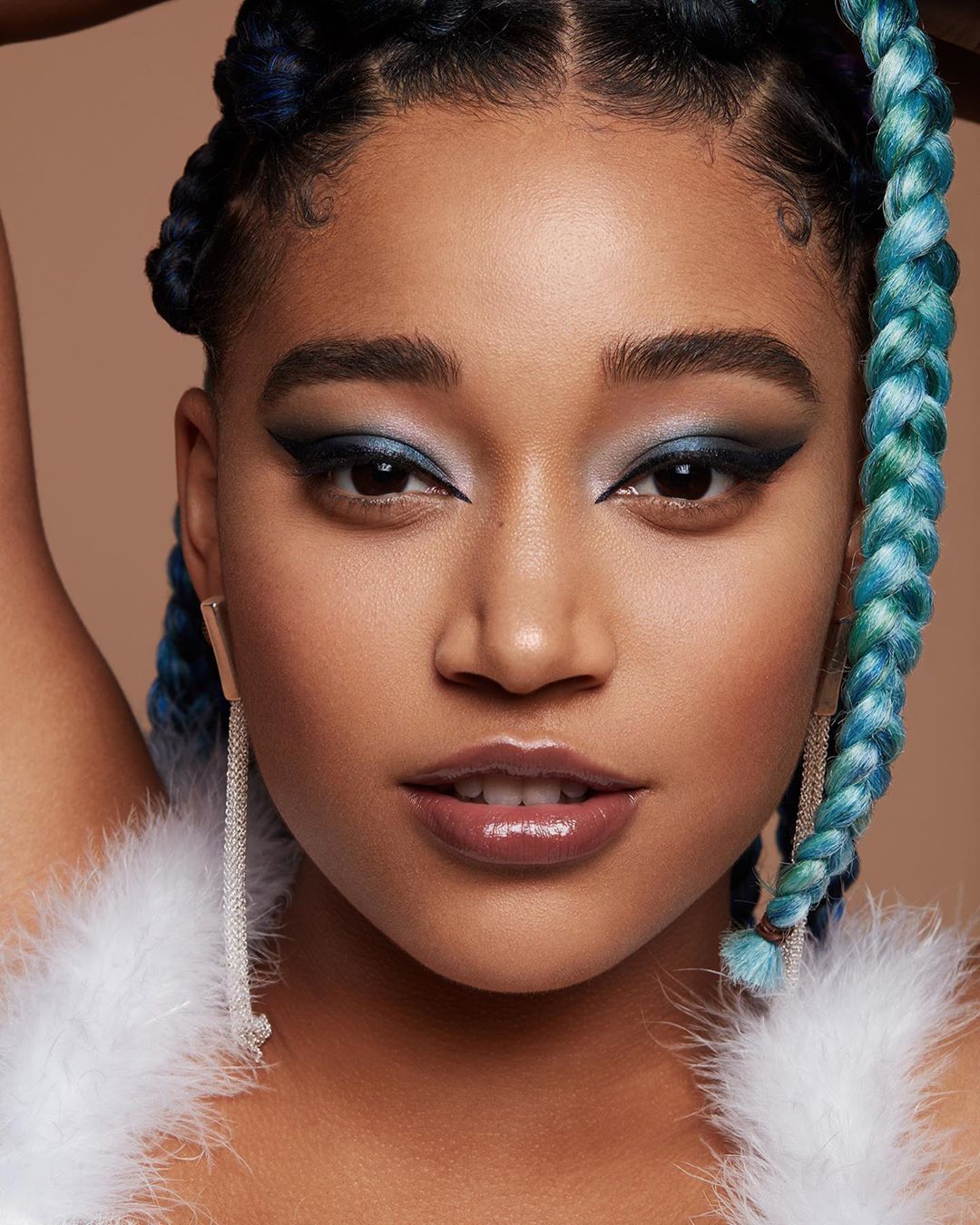 Close-up portrait of a woman with blue braided hair, wearing dramatic blue eye makeup and glossy lips against a tan background.