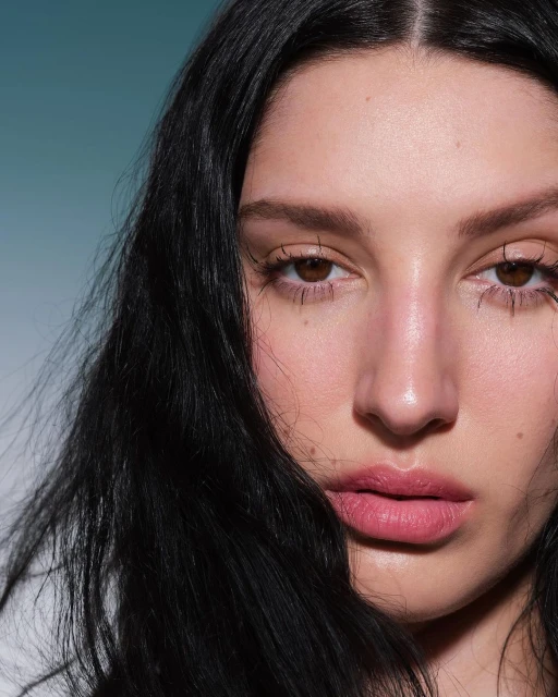Close-up portrait of a young adult woman with long, dark hair and light skin against a soft blue background in a beauty shot.