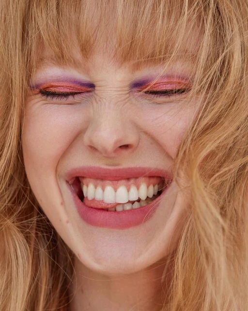Close-up portrait of a young woman with closed eyes and vibrant makeup, smiling widely and sticking out her tongue. Light blonde hair frames her face.