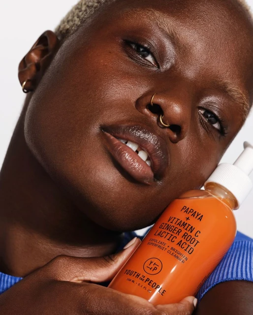 Close-up of a dark-skinned person with short blonde hair holding an orange bottle of Youth to the People Papaya + Vitamin C cleanser.