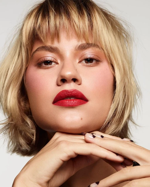 Close-up of a young woman with blonde bob and bangs, red lipstick, and manicured nails posing against a neutral background.