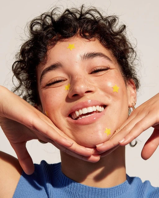 Smiling young woman with curly hair and fair skin, several yellow star stickers on her face, framing her face with her hands.