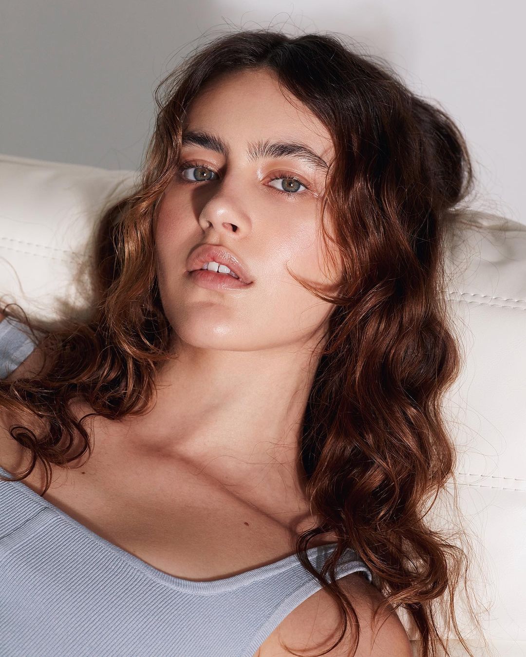 Close-up portrait of a woman with long wavy brown hair wearing a light blue ribbed top against a white surface