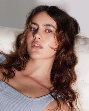Close-up portrait of a woman with long wavy brown hair wearing a light blue ribbed top against a white surface