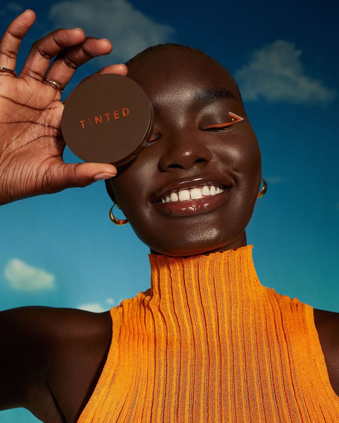 Smiling woman with dark skin holds brown tinted foundation compact. She wears an orange top and gold hoops against a cloudy sky.